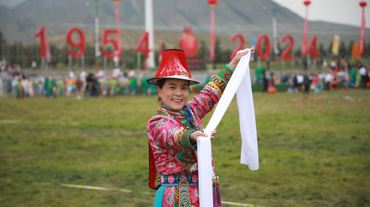 Yugur woman in colorful traditional clothing holding a white ceremonial scarf at an outdoor celebration