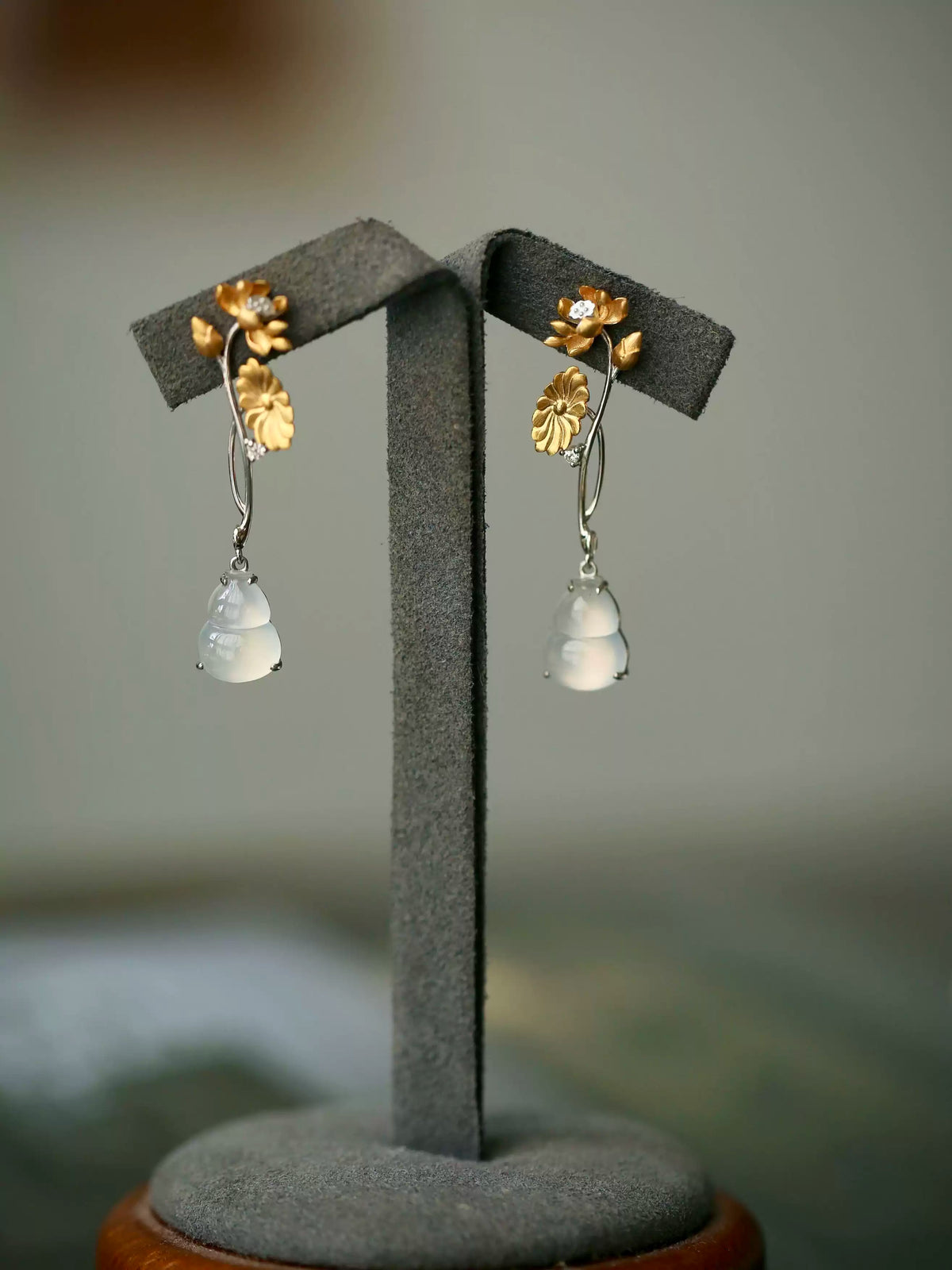 Pair of long earrings with gold-tone flower details and pale imitation-jade-style gourd drops, arranged on a display stand.