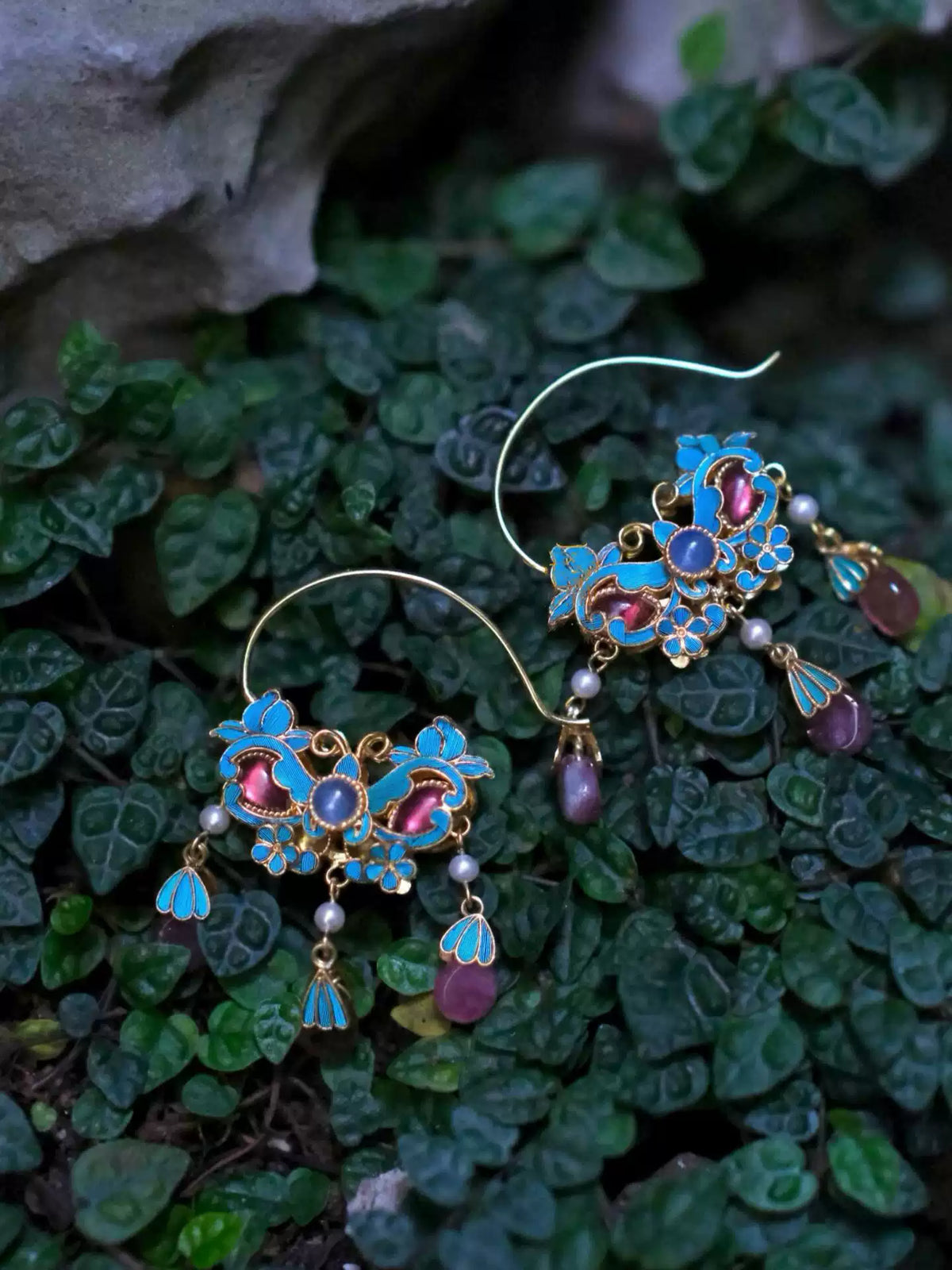 Pair of blue enamel floral tassel earrings displayed against green leaves.