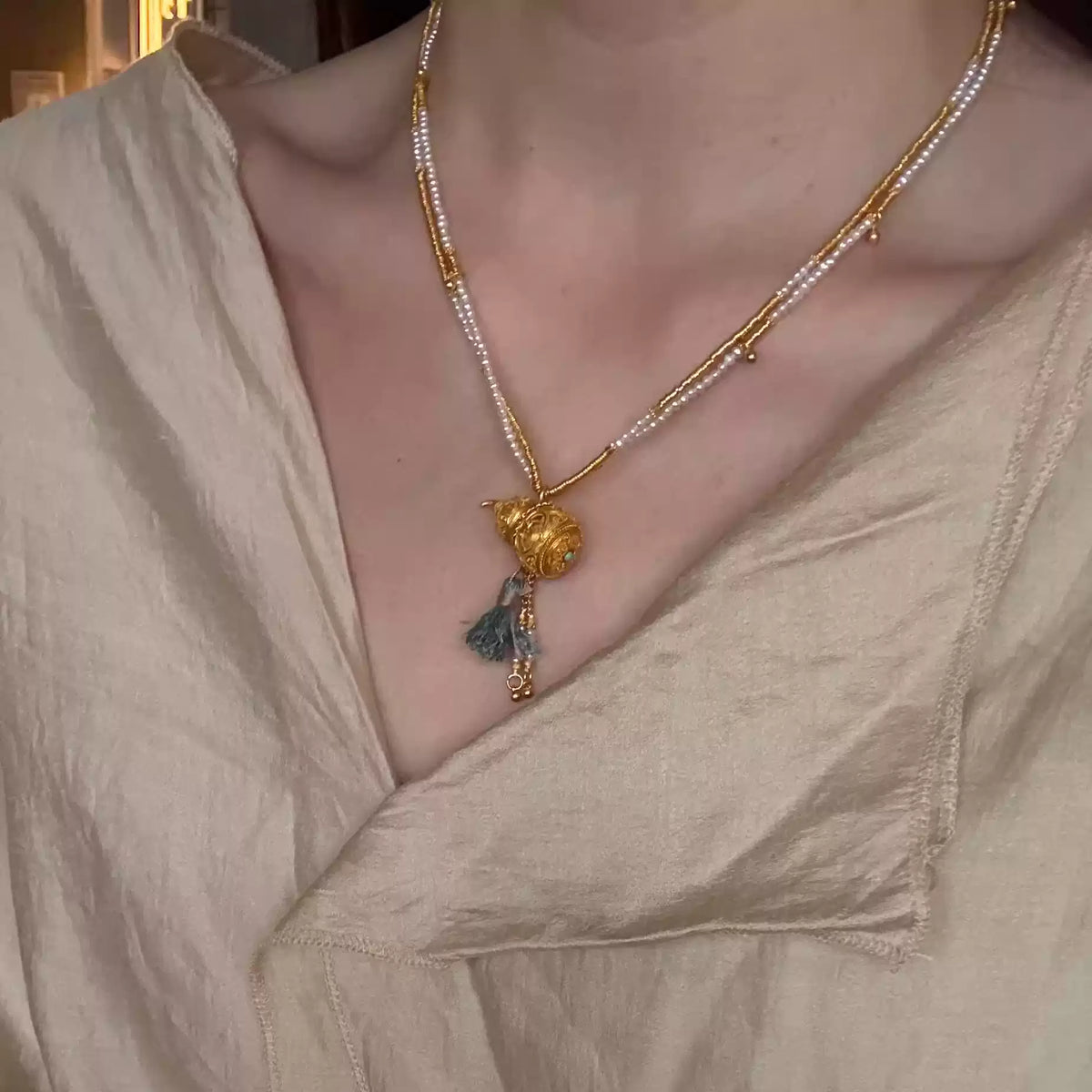 Gold-tone necklace with gourd pendant and blue-green tassel, worn with a light outfit