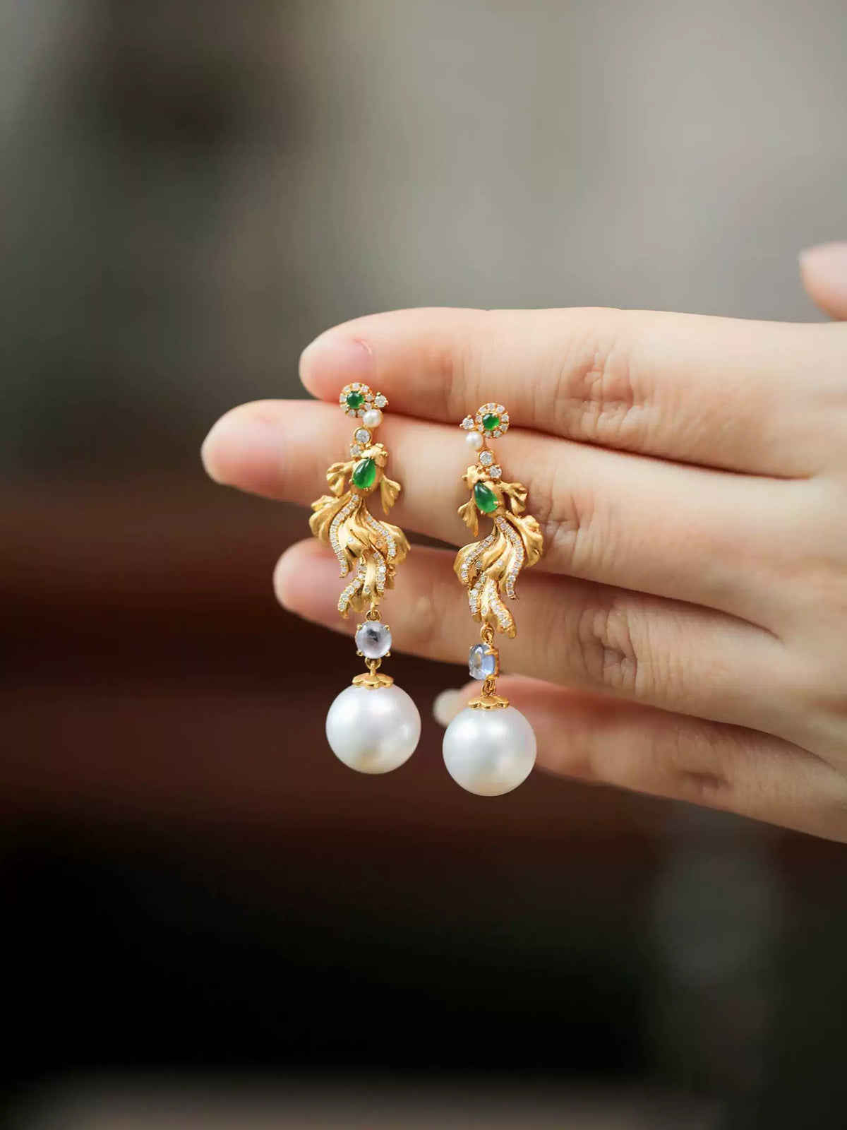 Pair of gold-tone koi fish drop earrings with green imitation jade accents and white round drops, worn on hand