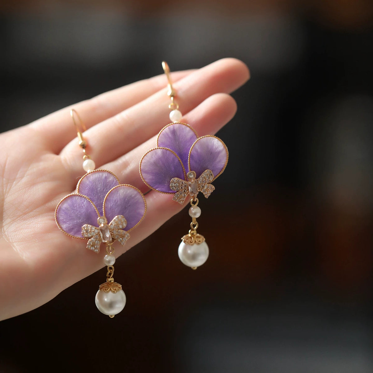 Pair of violet velvet flower drop earrings with white bead drops, displayed on a hand